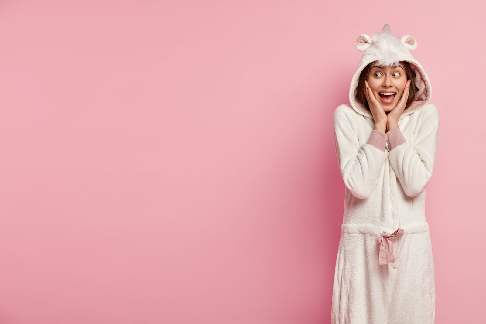 Image of positive woman touches cheeks, enjoys good morning, smiles positively, looks away on free space, dressed in soft domestic kigurumi costume, isolated on pink background. Sleeping concept